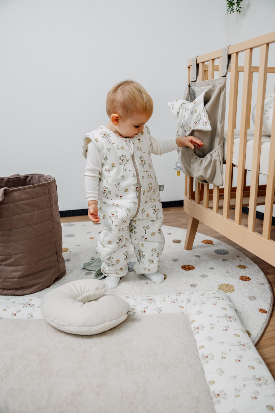Aktives Kind spielt im Kinderzimmer im Odenwälder Winter Jersey-Hopsi Babyschlafsack, creme mit Bäumchen, atmungsaktiv und weich, bequem und flexibel für Kinder, die schon flitzen und sich viel bewegen. Neben ihm liegt auf dem Boden das Nackenhörnchen für unterwegs.