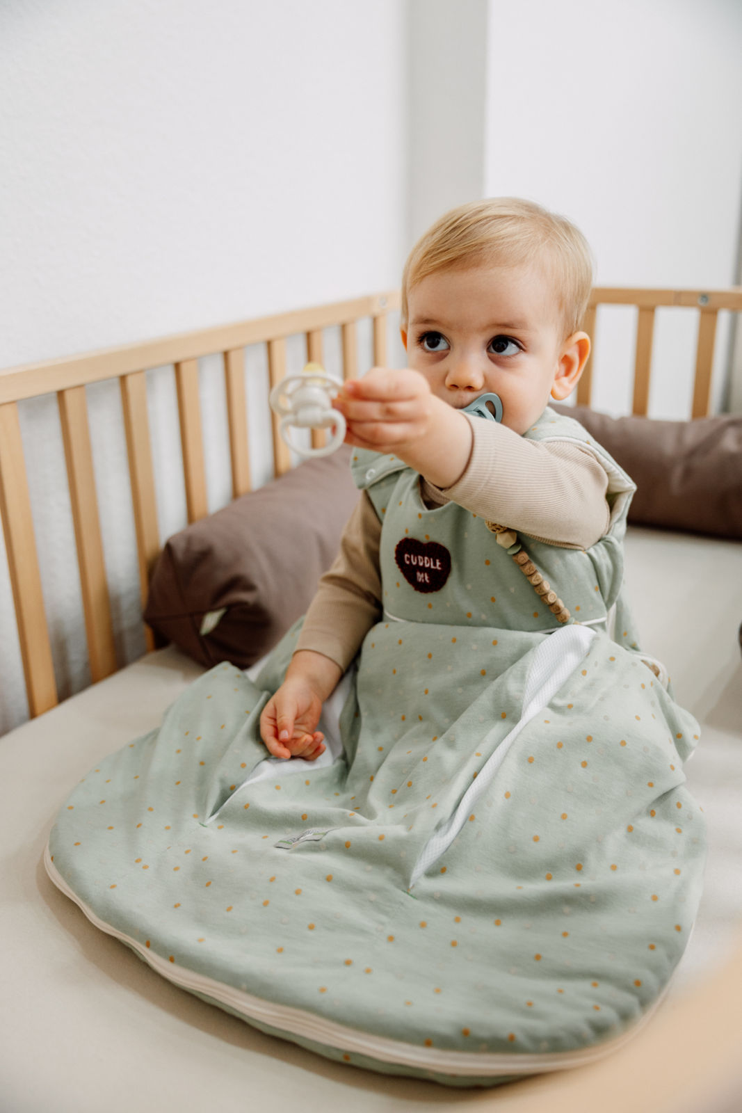 Baby trägt einen prima klima Jersey‑Schlafsack in der Designvariante Dotty mit gepunktetem Muster, ärmellos, aus weichem Jersey-Stoff, in natürlicher Umgebung sitzend im Babybett, hält einen Schnuller in der Hand.
